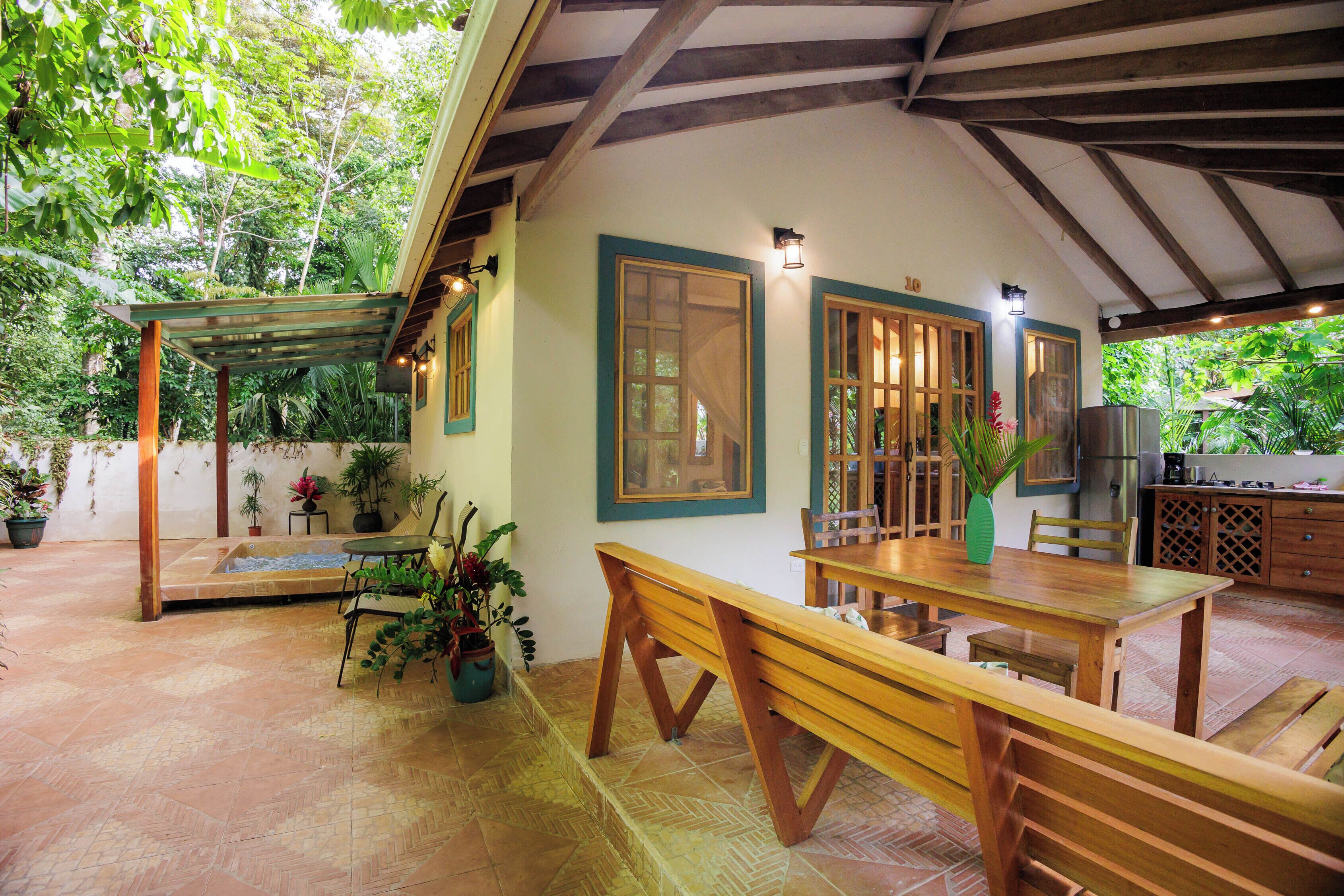 An outdoor patio with a wooden table and benches, surrounded by potted plants and a pergola.