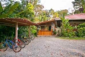 A building with a gravel driveway, a covered bike parking area, and lush greenery around. - Casa Merlin Lodge (Cahuita)