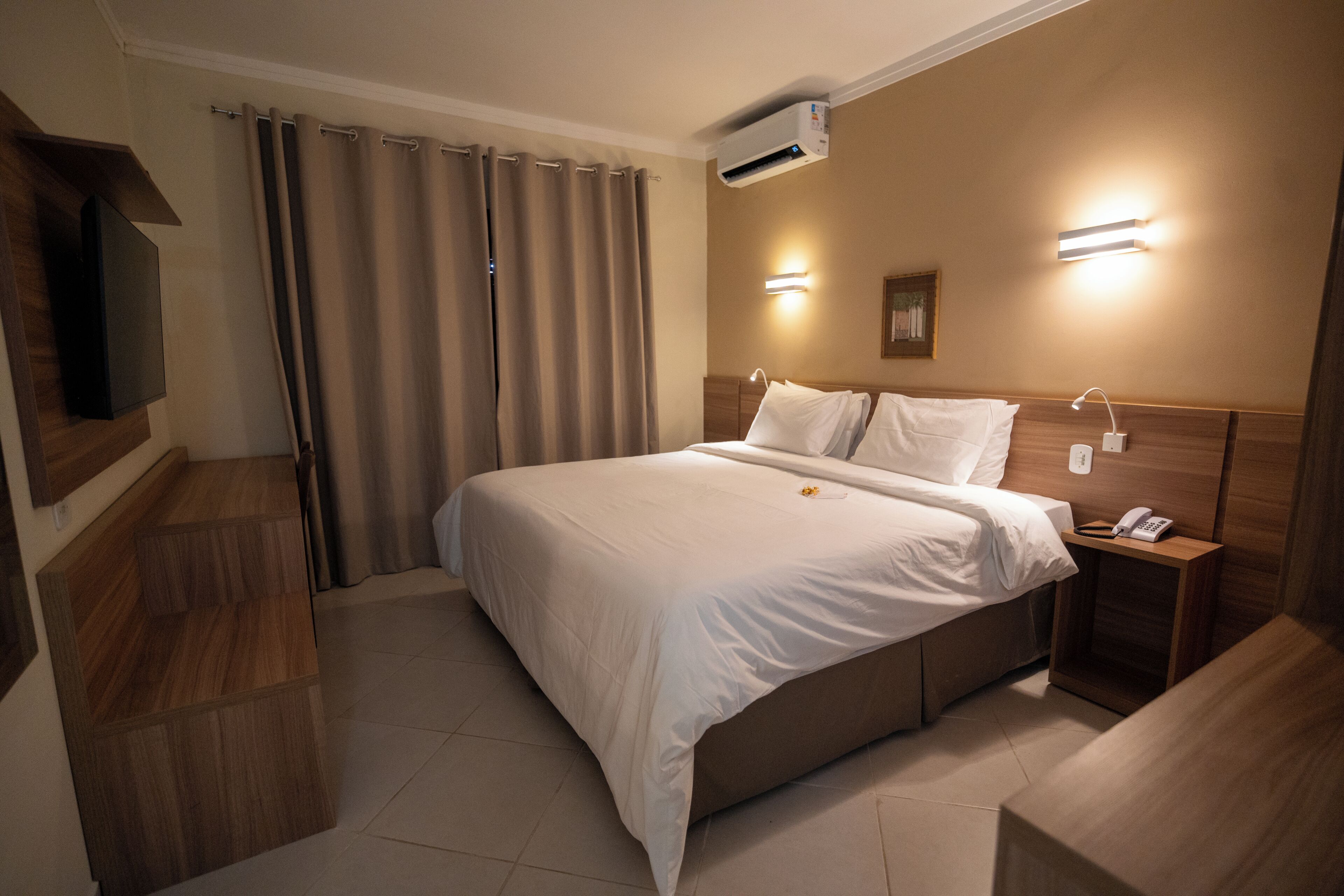 A hotel room with a bed, bedside tables, an air conditioning unit, and a television.
