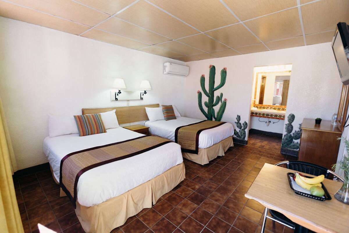 A hotel room with two beds, a desk with a fruit basket, and a cactus mural on the wall.