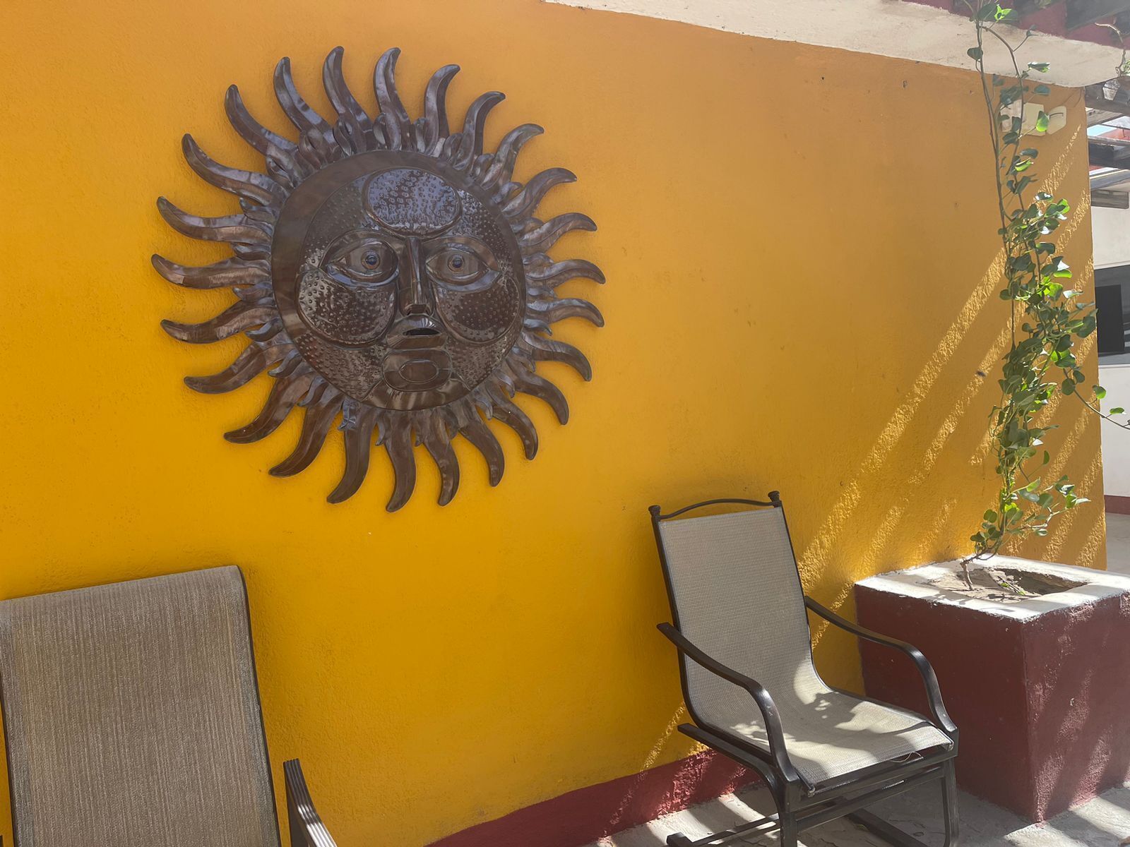 A yellow wall with a metal sun sculpture, a metal chair, and a concrete planter with a plant.