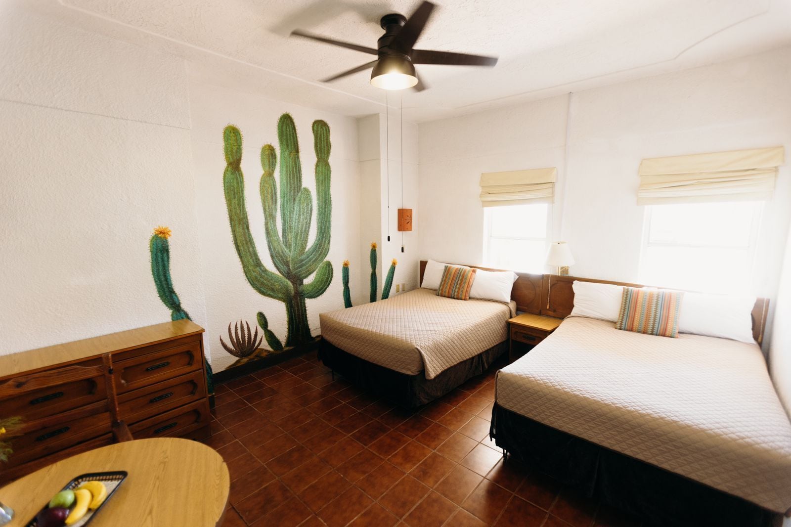 A room with two beds, a wooden dresser, a round table with fruit, and a cactus mural on the wall.