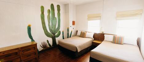 A room with two beds, a wooden dresser, a round table with fruit, and a cactus mural on the wall.