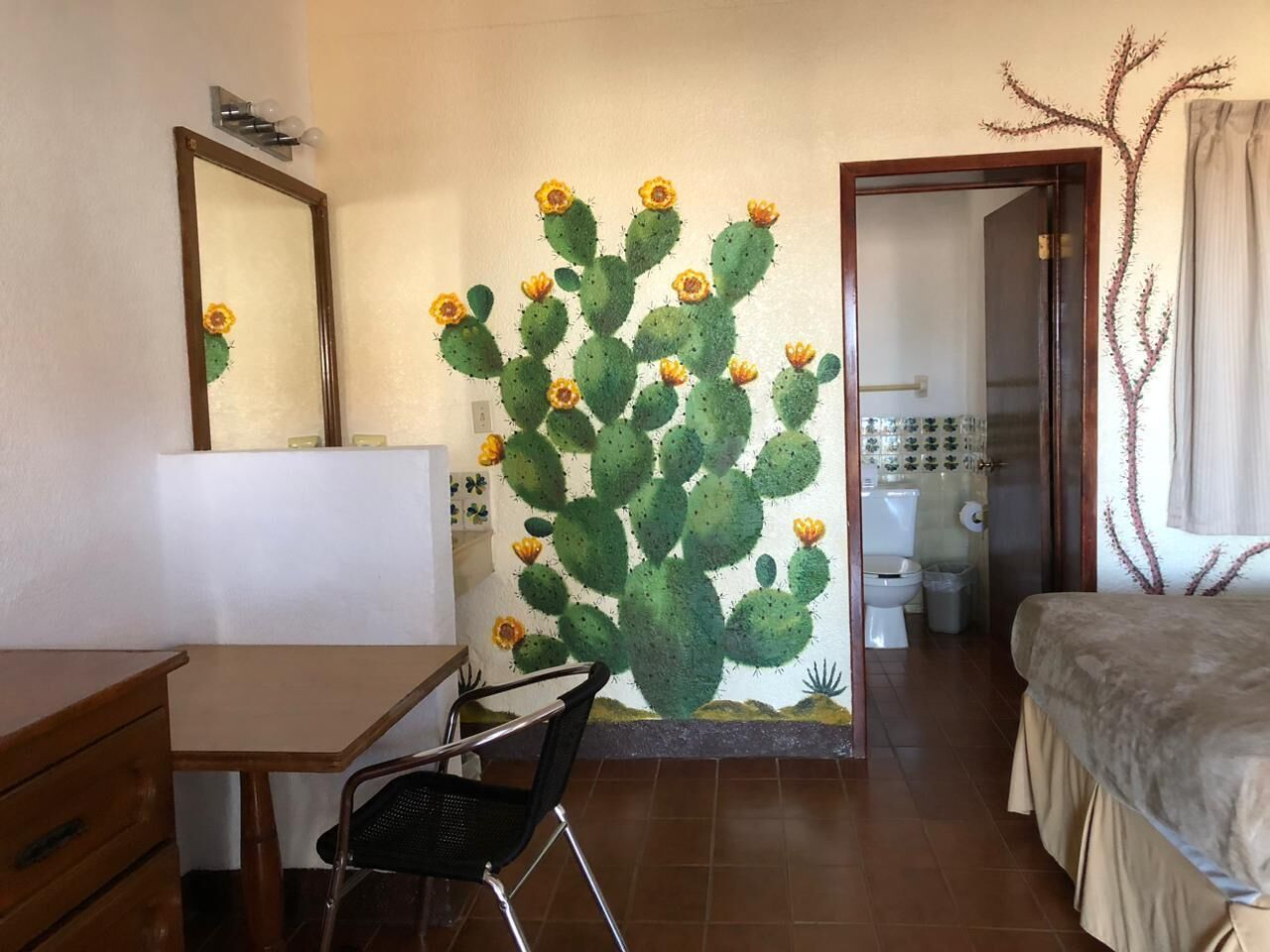 A room with a cactus mural, a desk, a chair, a bed, and a bathroom visible through an open door.
