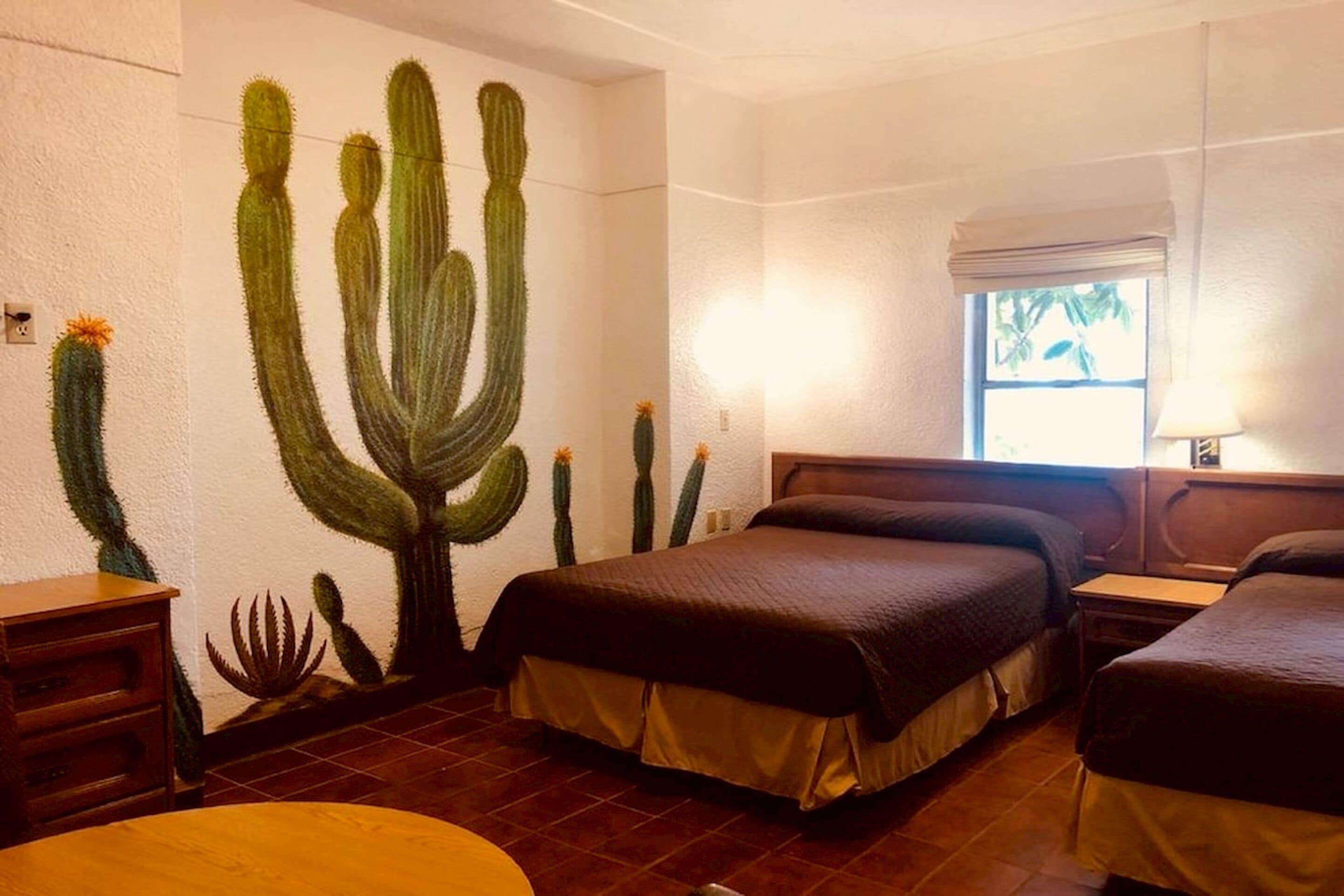 A hotel room with two beds, a cactus mural, a window with a view, and a wooden nightstand.