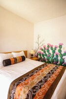 Habitación de hotel con cama, mesita de noche, lámpara y un mural de cactus en la pared.