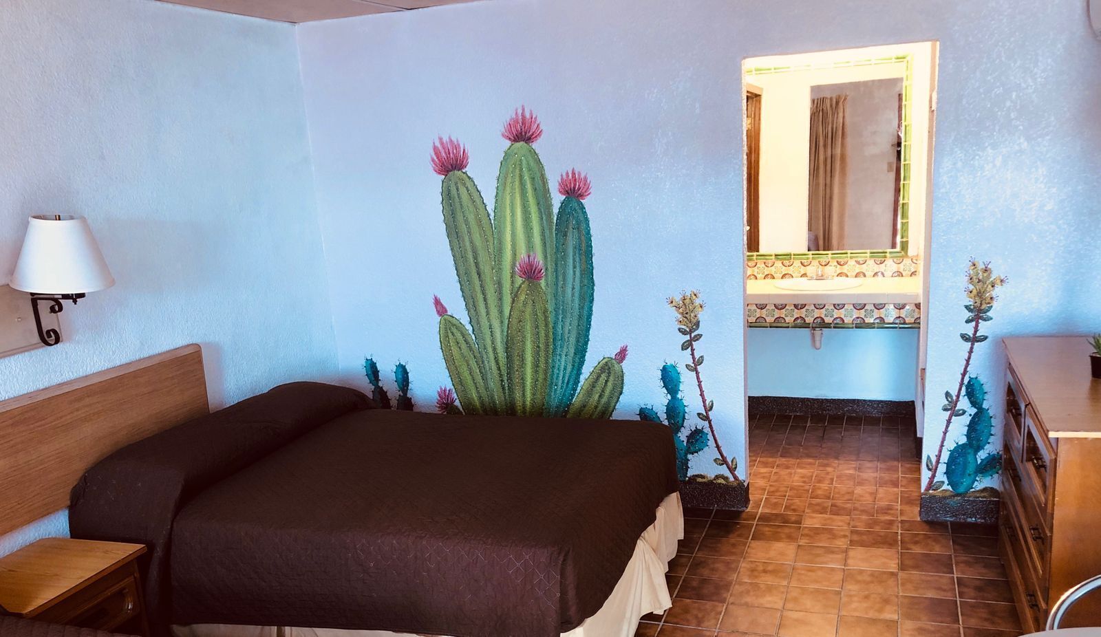 A bedroom with a bed, a nightstand, a lamp, a mirror, and a wall mural of cacti.