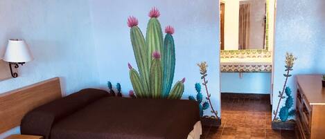 A bedroom with a bed, a nightstand, a lamp, a mirror, and a wall mural of cacti.