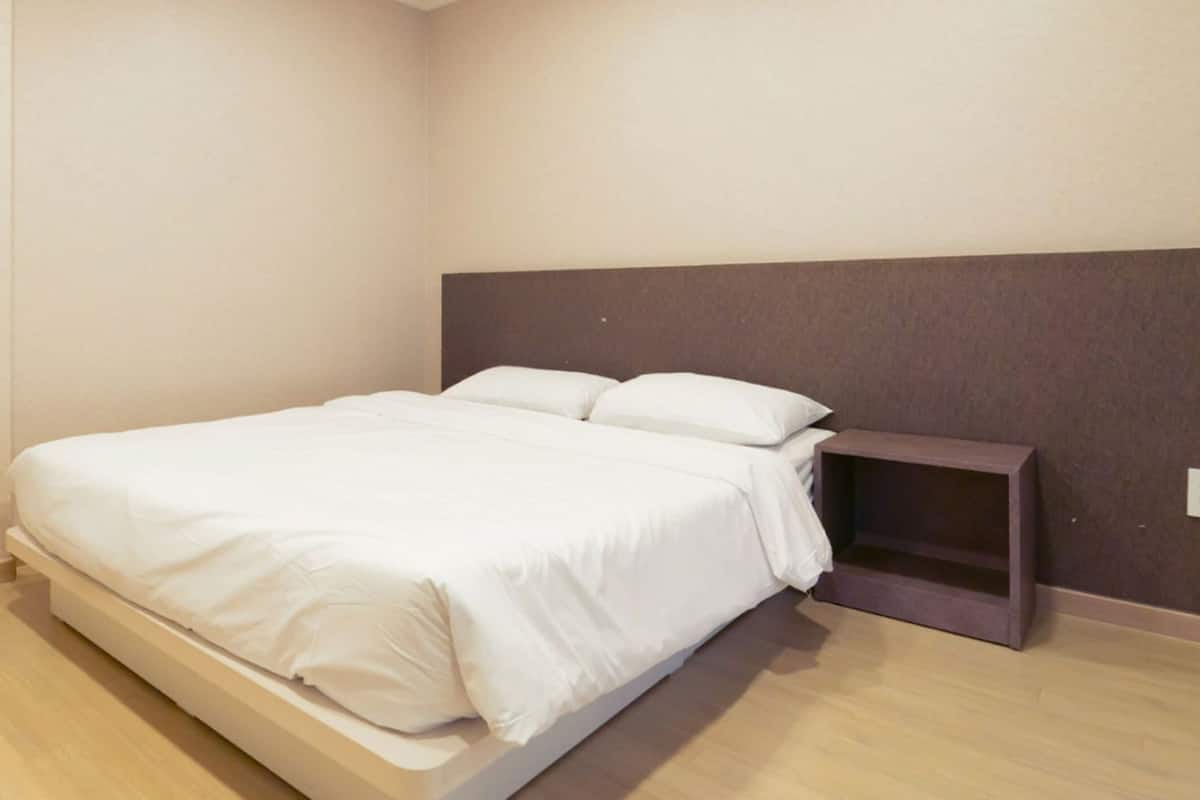 Una cama individual con ropa de cama blanca en una habitación con suelo de madera clara y cabecera oscura.