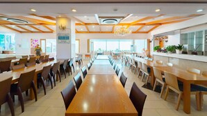 Un vaste espace dédié à la salle à manger, avec de longues tables et des chaises en bois, un lustre central et une vue sur l’extérieur grâce à de grandes fenêtres.