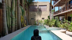 Un espace piscine paisible, avec une statue de Bouddha au premier plan, entouré d’une végétation tropicale luxuriante et, au fond, un bâtiment doté de balcons.
