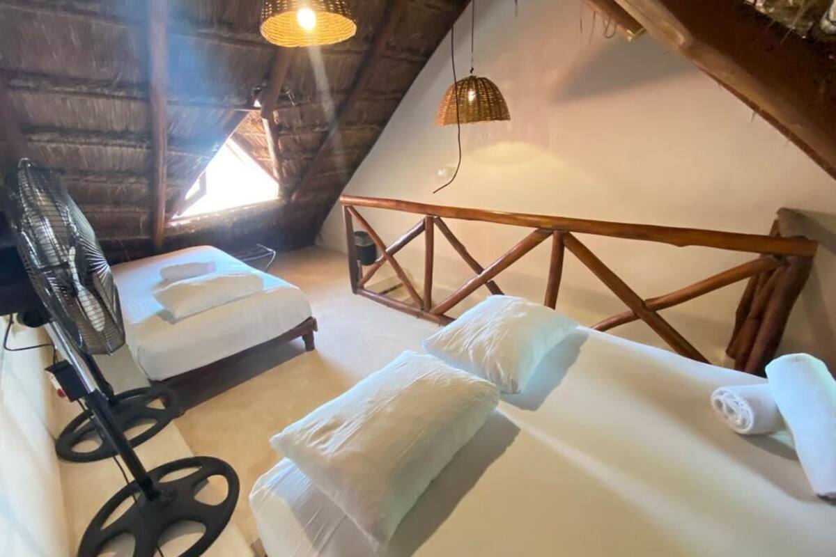A cozy attic room with two beds, a fan, and a woven light fixture.