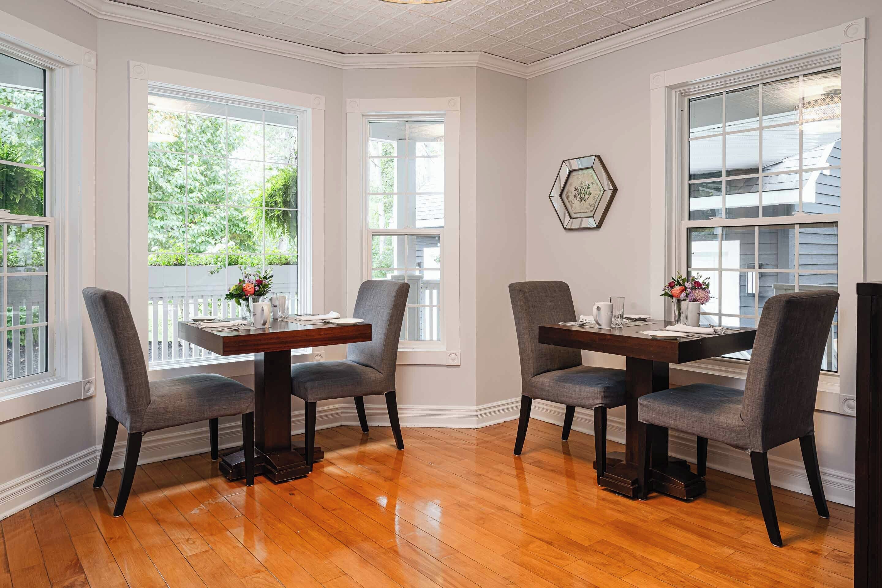 A dining area with a wooden table and four chairs, large windows with a view of greenery, and a decorative hexagonal mirror on the wall.