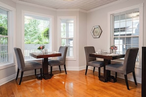 Un comedor con una mesa de madera y cuatro sillas, ventanas amplias con vistas a follaje, y un espejo hexagonal decorativo en la pared.
