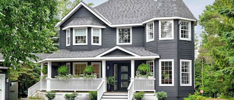 A two-story house with a steeply pitched roof, a front porch with white railings, and a dark grey siding exterior.