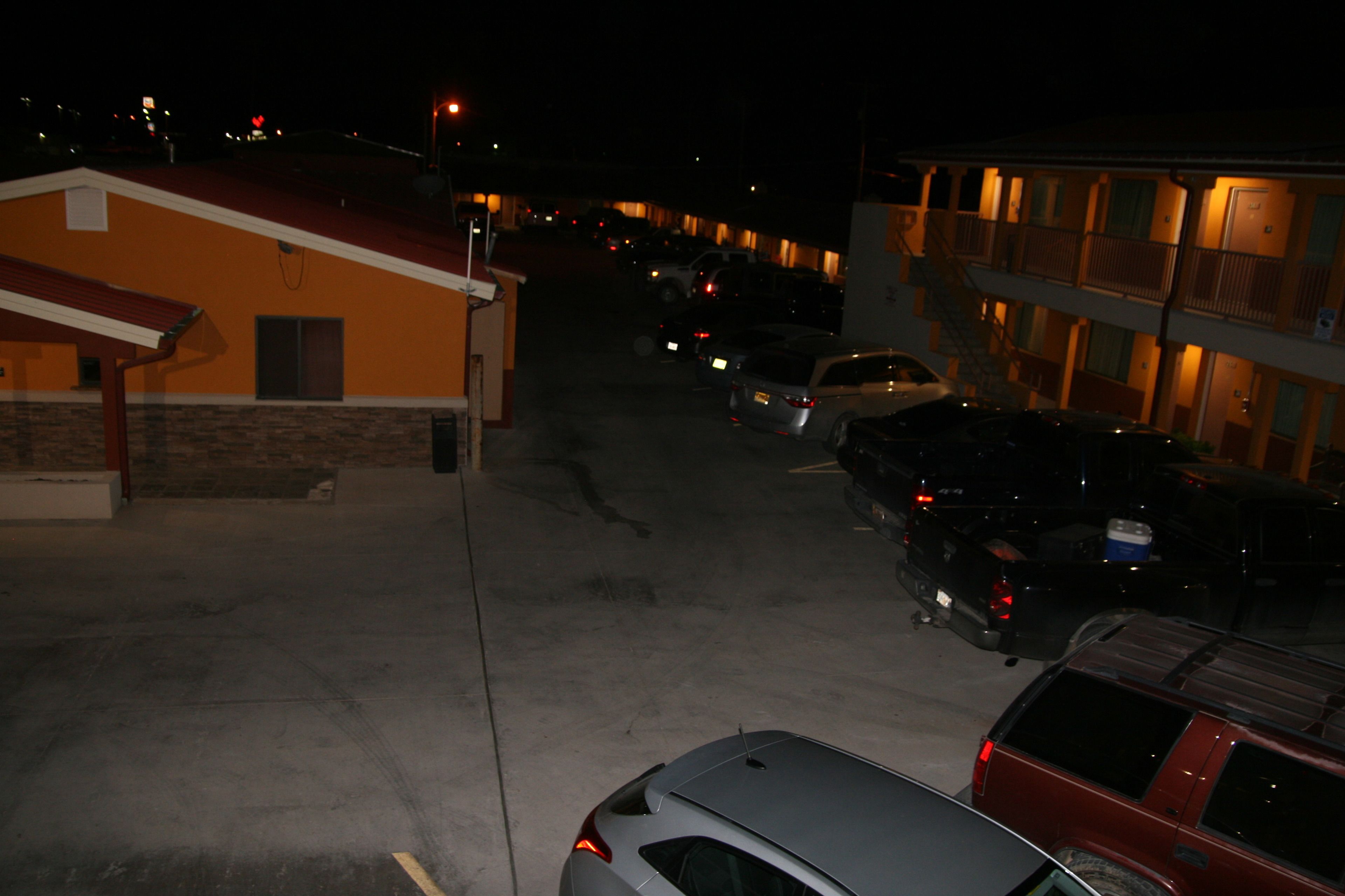 A parking lot with several cars parked in front of a building with a red roof and a balcony on the second floor.