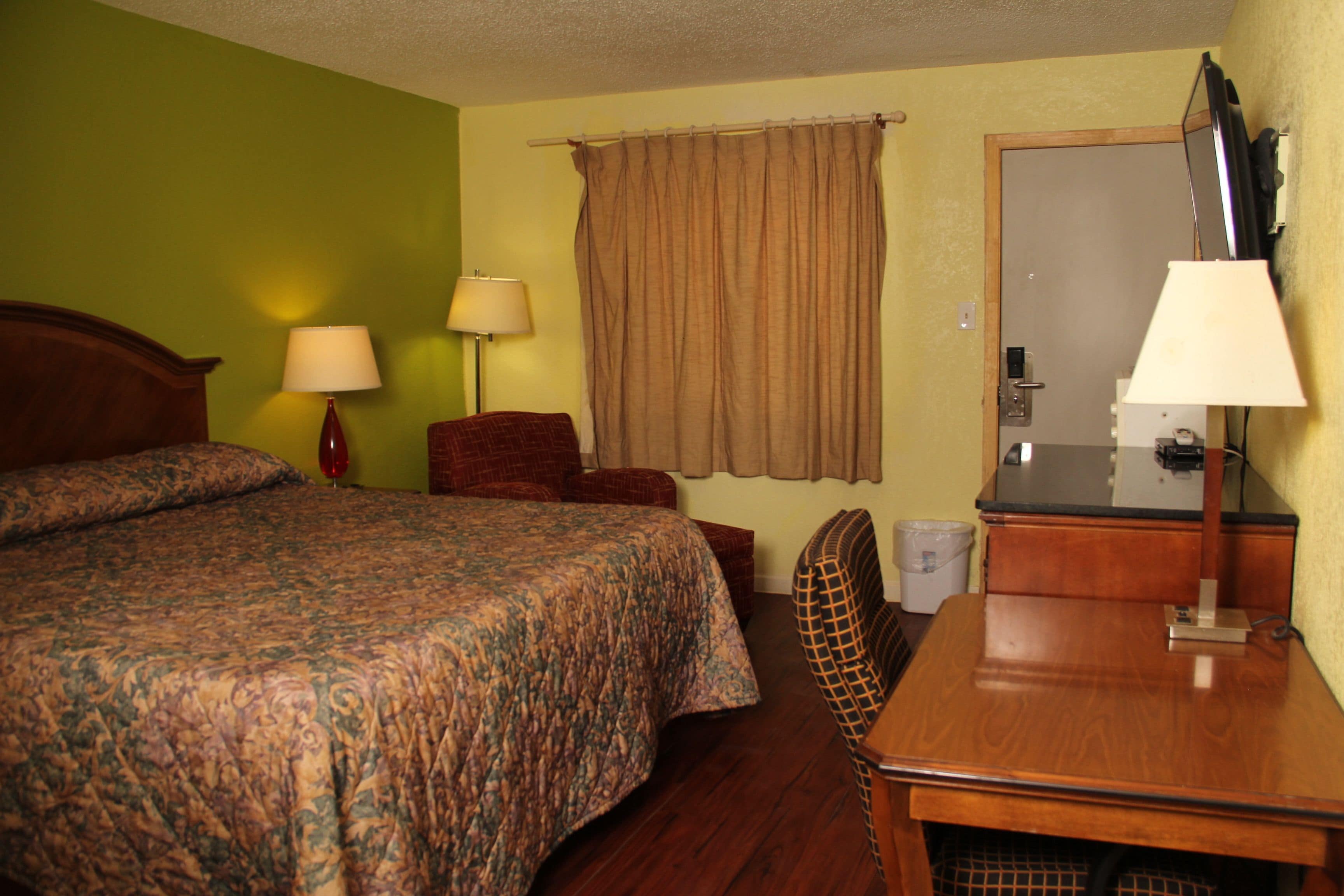 A hotel room with a bed, a desk, a chair, a lamp, and a curtain.