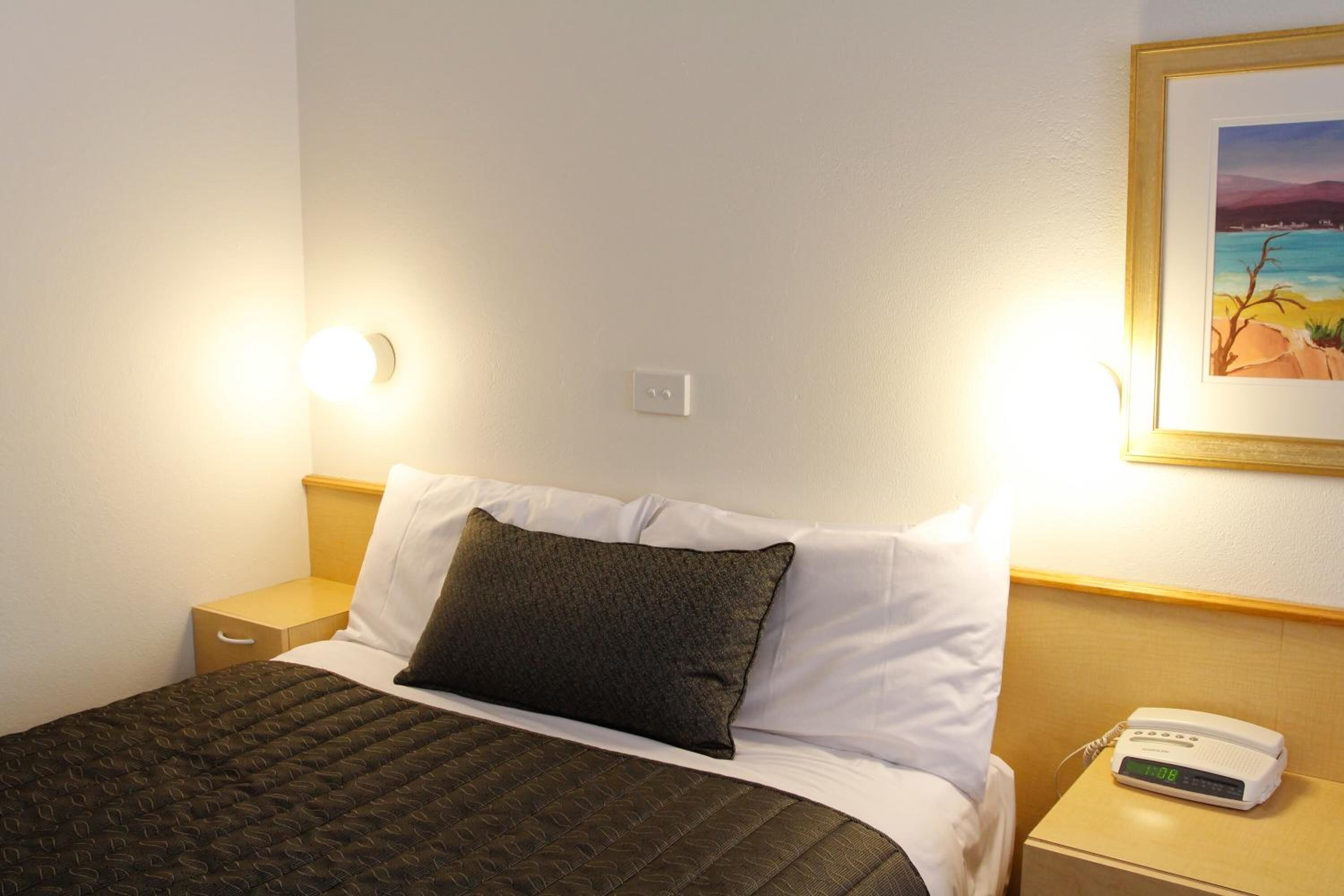 A hotel room with a bed, a nightstand, a digital clock, and a framed picture on the wall.