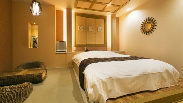 A neatly arranged hotel room with a bed, two wicker chairs, a mirror, and a sun-shaped wall decoration. - Hotel Atlantis Higashiosaka - Adults Only (Higashi-osaka)