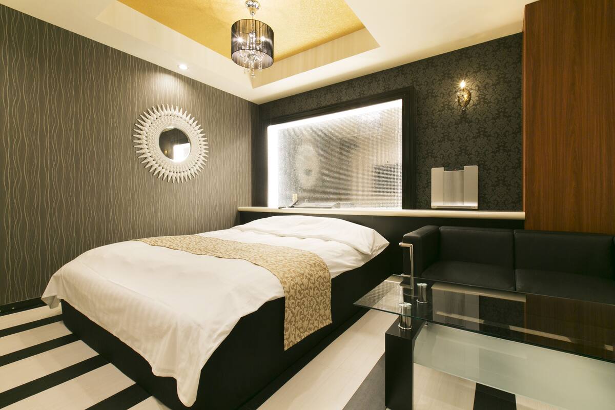A modern bedroom with a large bed, a glass coffee table, a black sofa, a round mirror, and a large window with blinds.