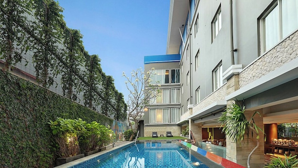A swimming pool with tiled flooring, surrounded by greenery and a building with a stone facade. - Primera Hotel Seminyak (Seminyak)