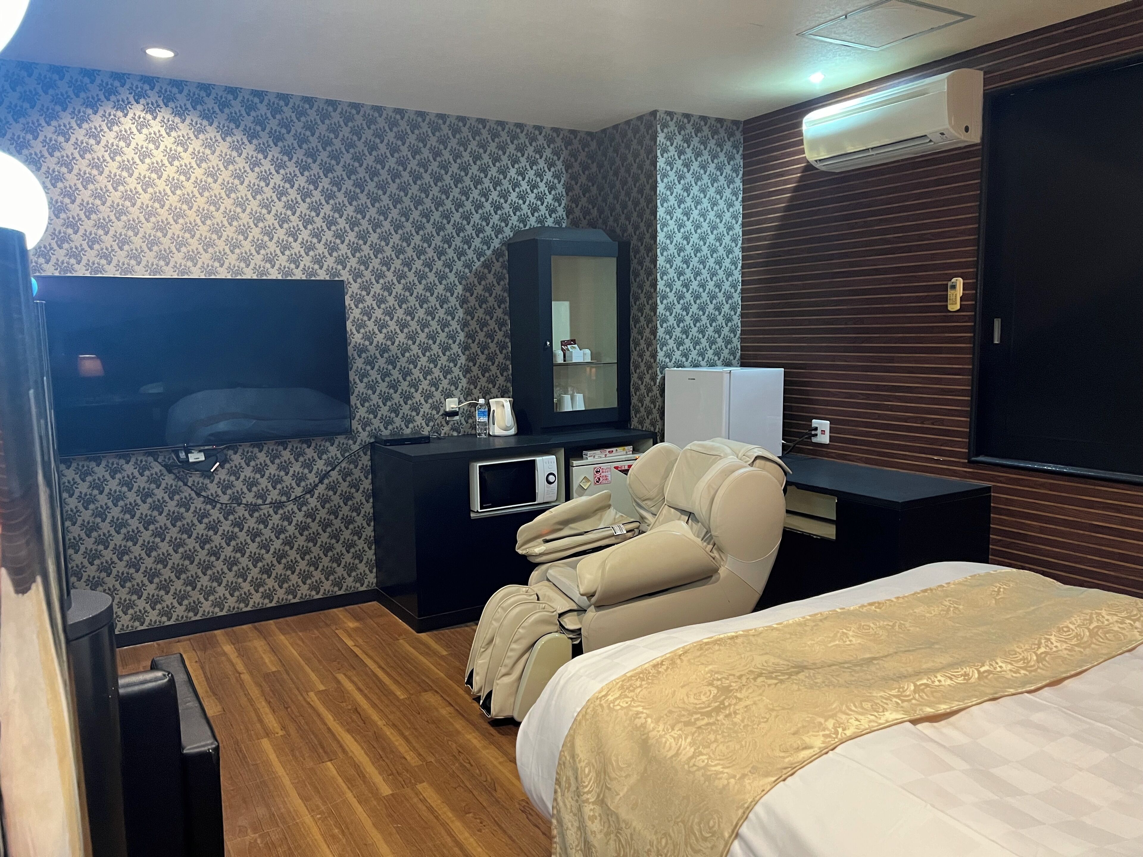 Une chambre d’hôtel équipée d’un lit, d’un fauteuil massant, d’un téléviseur à écran plat, d’un minibar et d’un climatiseur fixé au mur.