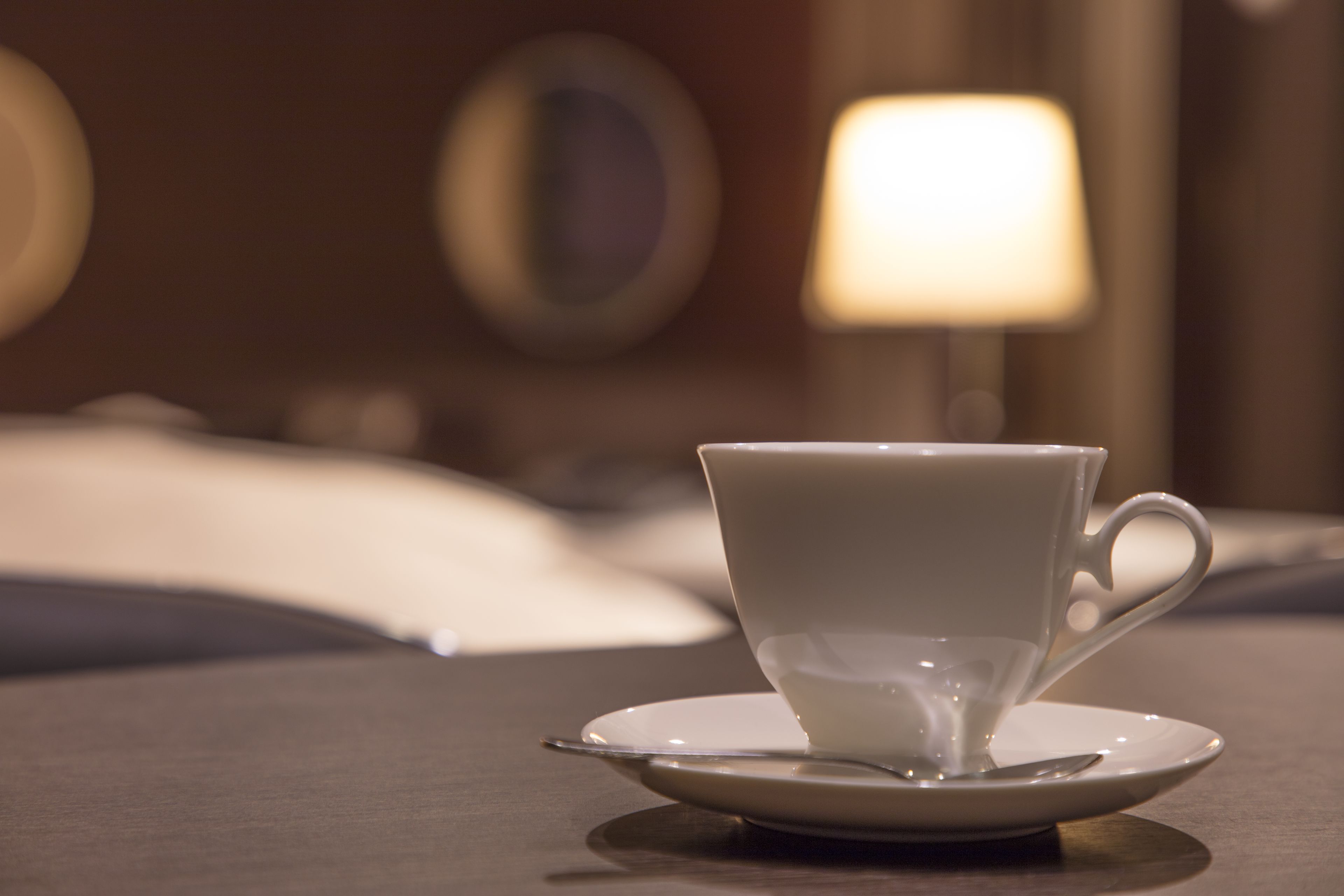 Une tasse posée sur une soucoupe, sur une table, dans une pièce où l’on aperçoit un lit et une lampe en arrière-plan.