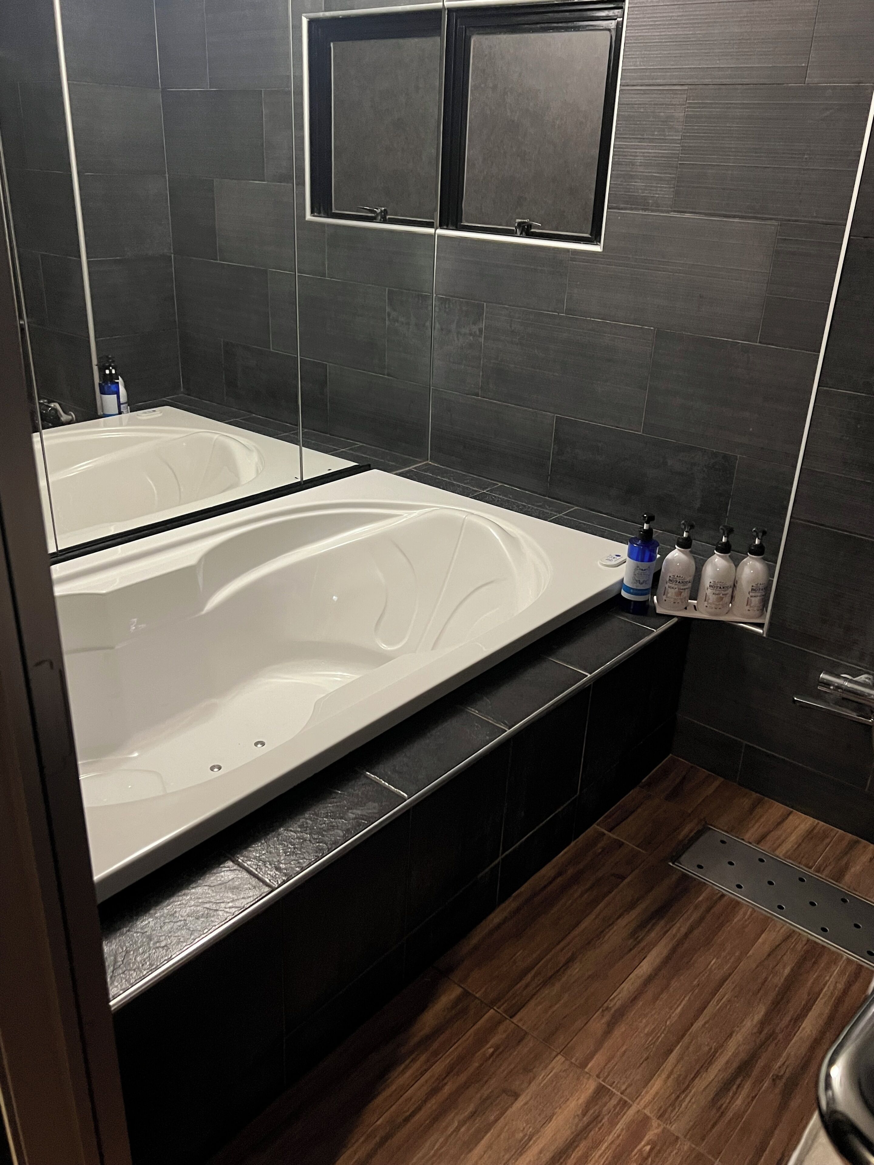 A modern bathroom with a large bathtub, dark tiled walls, and a wooden floor.