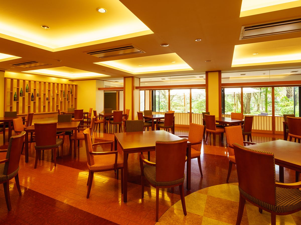 A spacious dining area with multiple tables and chairs, large windows offering a view of greenery outside, and a modern ceiling design with recessed lighting.