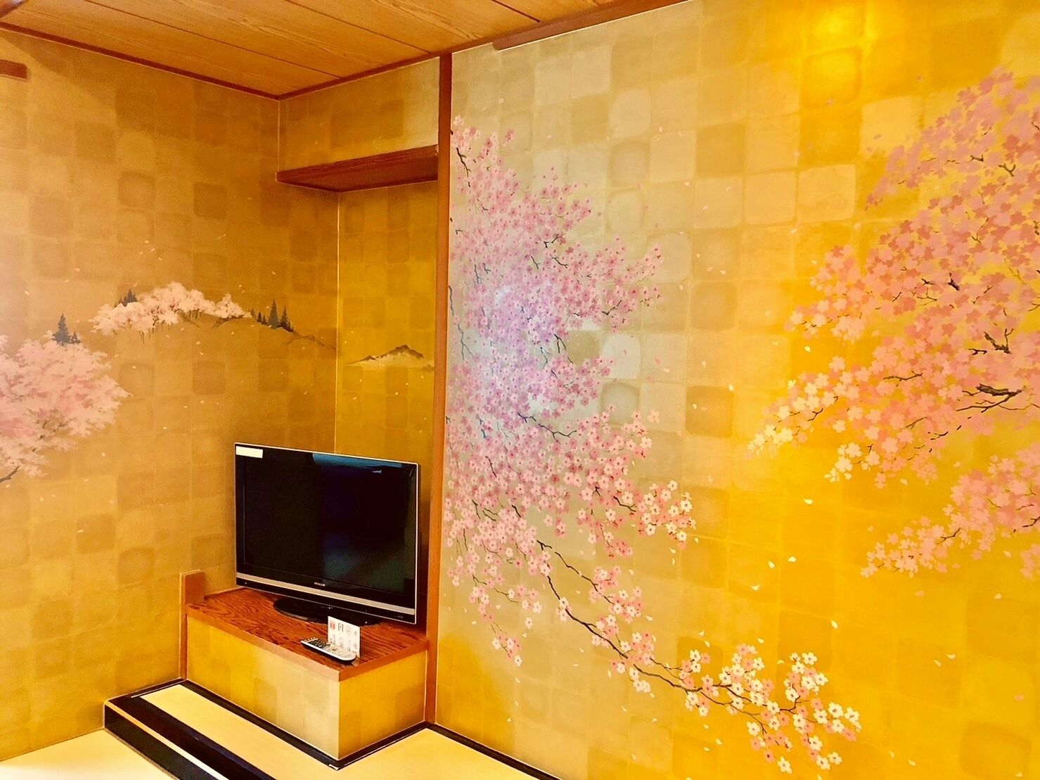 A room with a flat-screen TV on a wooden stand, a wall with a cherry blossom mural, and a tiled floor.