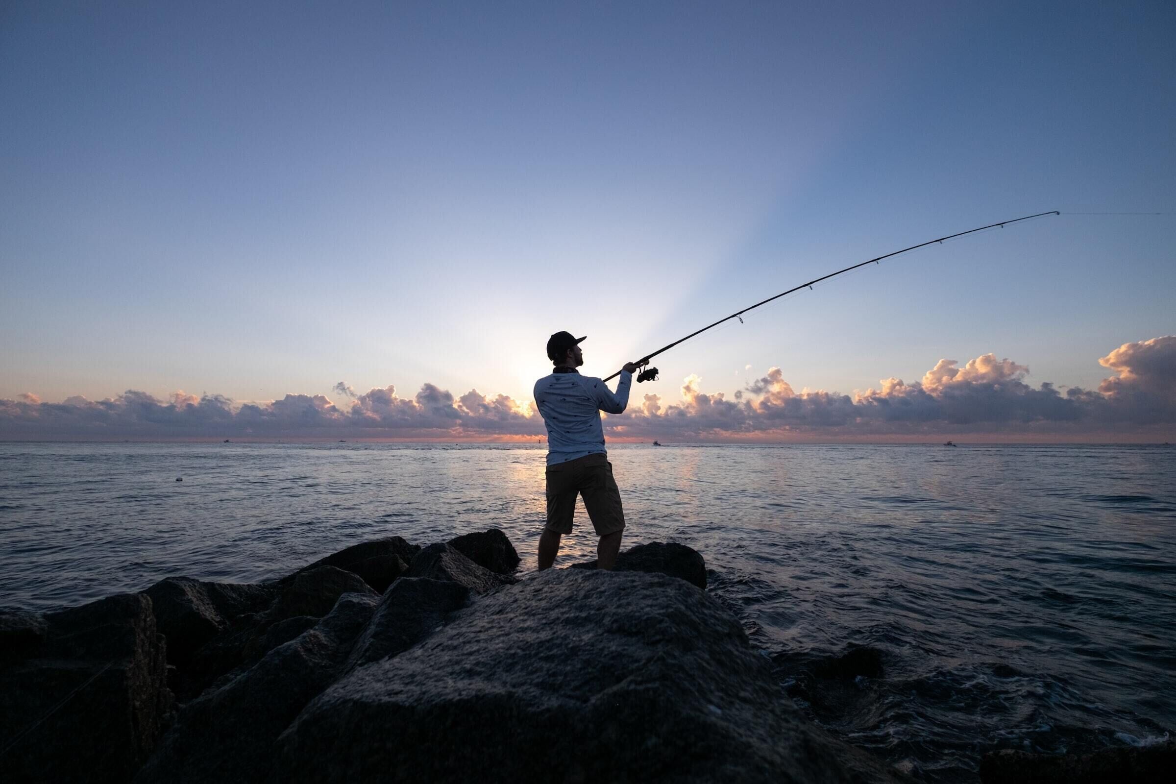 A person fishing at a rocky shoreline with a clear view of the ocean and sky.