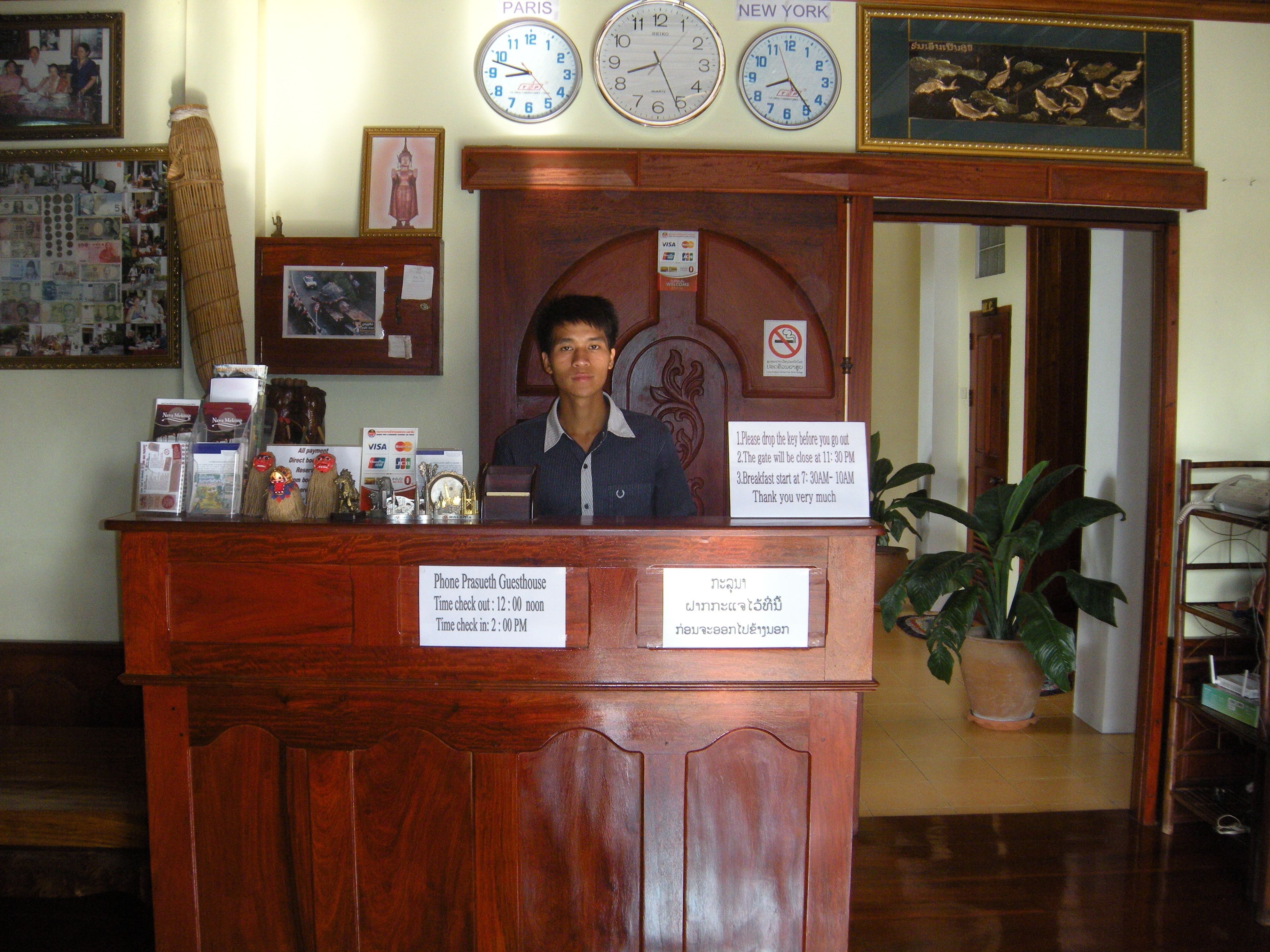 A wooden reception desk with clocks displaying Paris and New York times, a sign with operating hours, and a potted plant to the right.