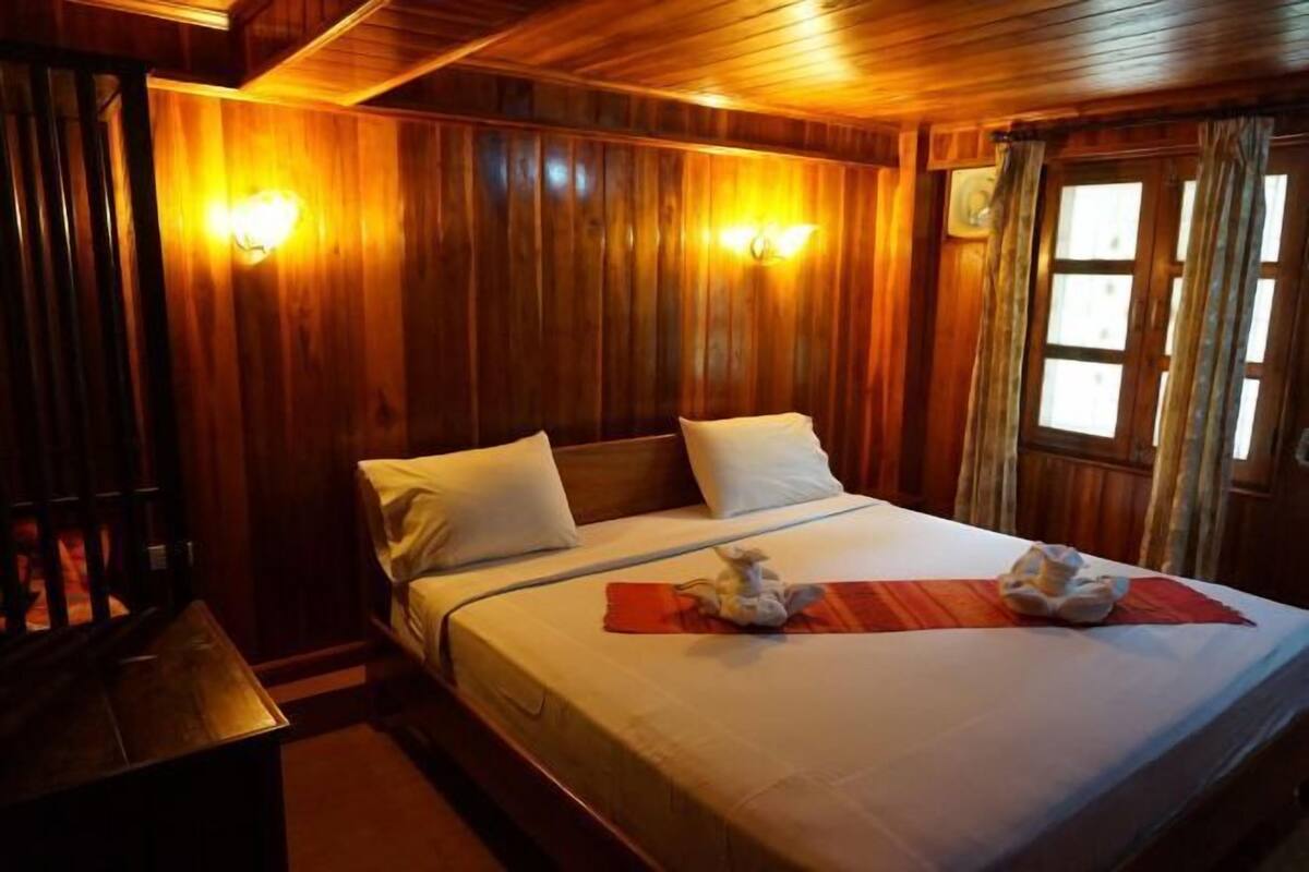 Ein Zimmer mit Holzwänden, einem Bett, zwei Wandleuchten und einem mit Vorhängen drapierten Fenster.
