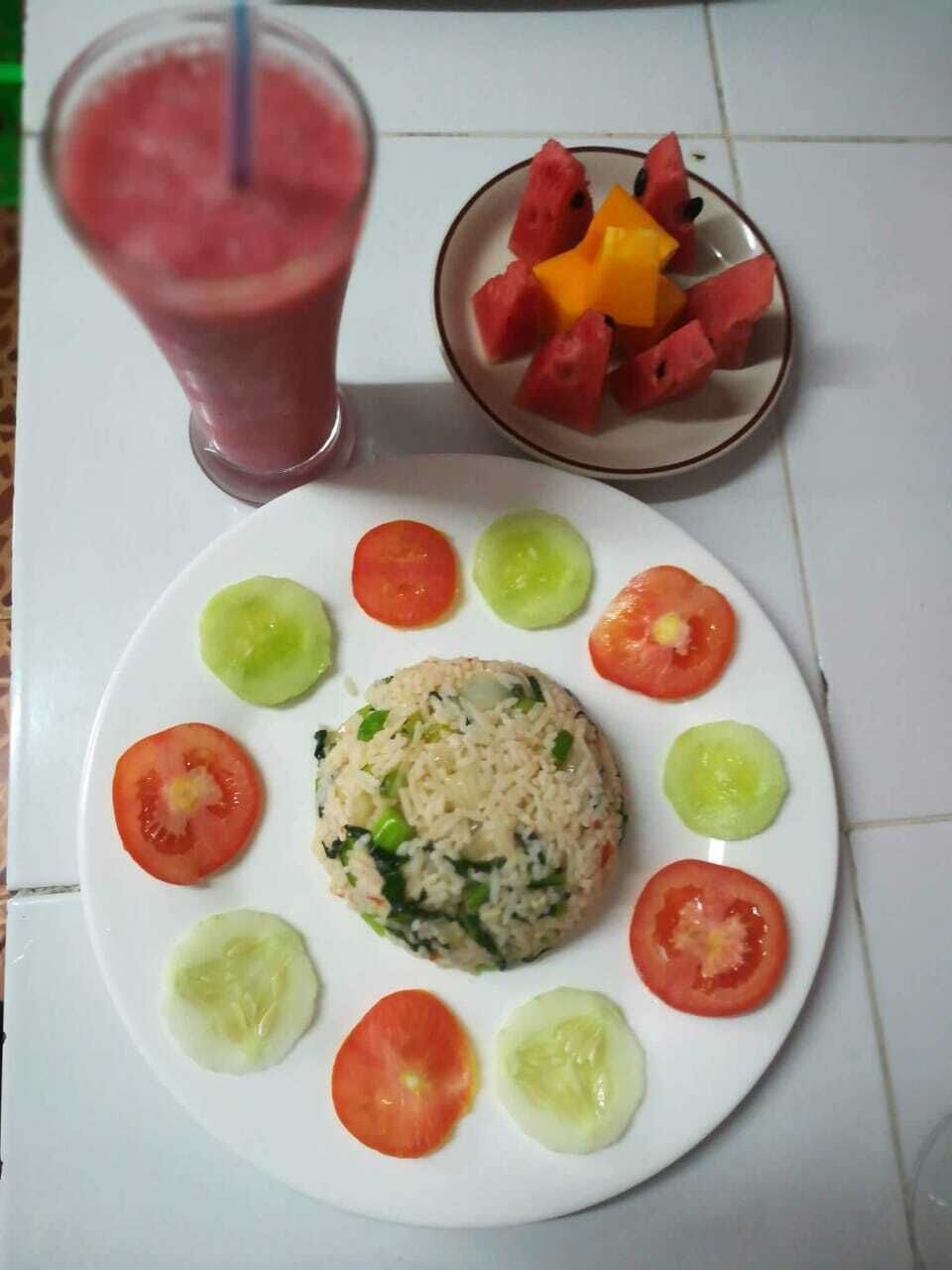 A plate with a rice-based dish surrounded by tomato slices, a glass of pink smoothie, and a bowl of watermelon pieces.