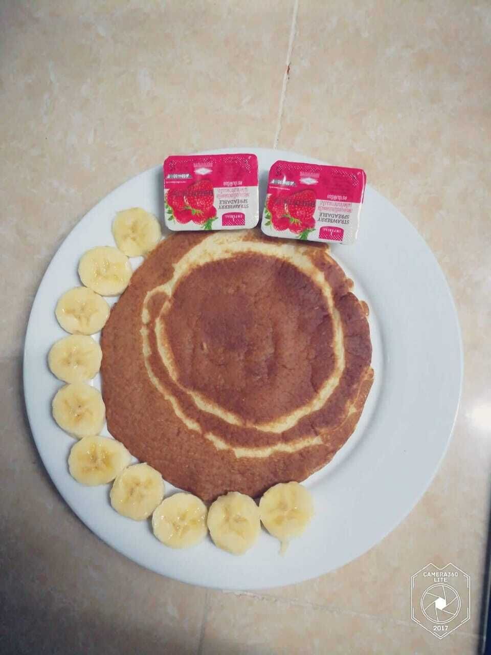 Een bord met een pannenkoek, plakjes banaan en twee bakjes aardbeienyoghurt.