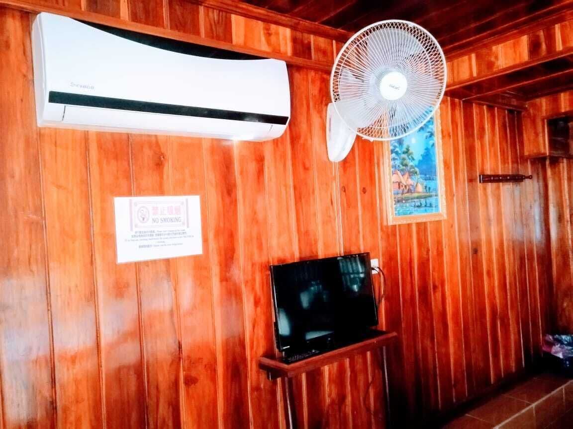 Una habitación con paneles de madera, un aire acondicionado blanco, un ventilador de pie, un televisor sobre un atril de madera y un cartel de 'No Fumar'.