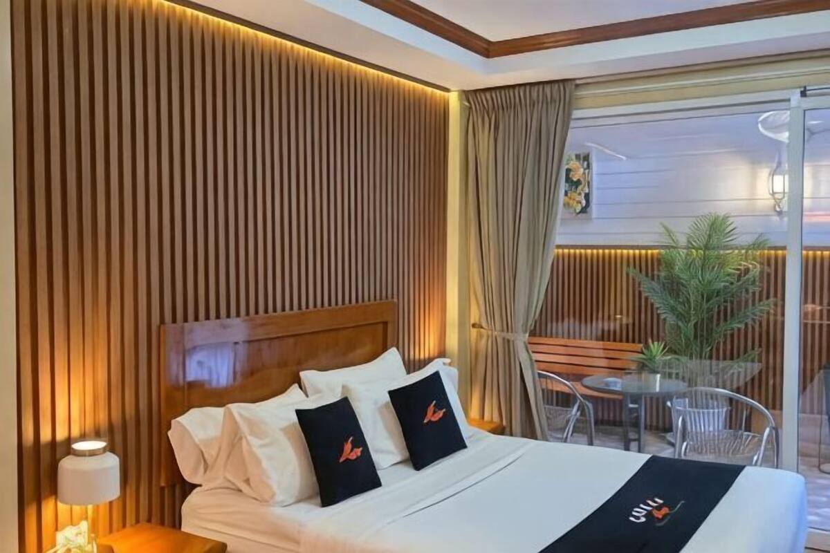 A hotel room with a bed, a nightstand, a lamp, a balcony with a table and chairs, and a potted plant.