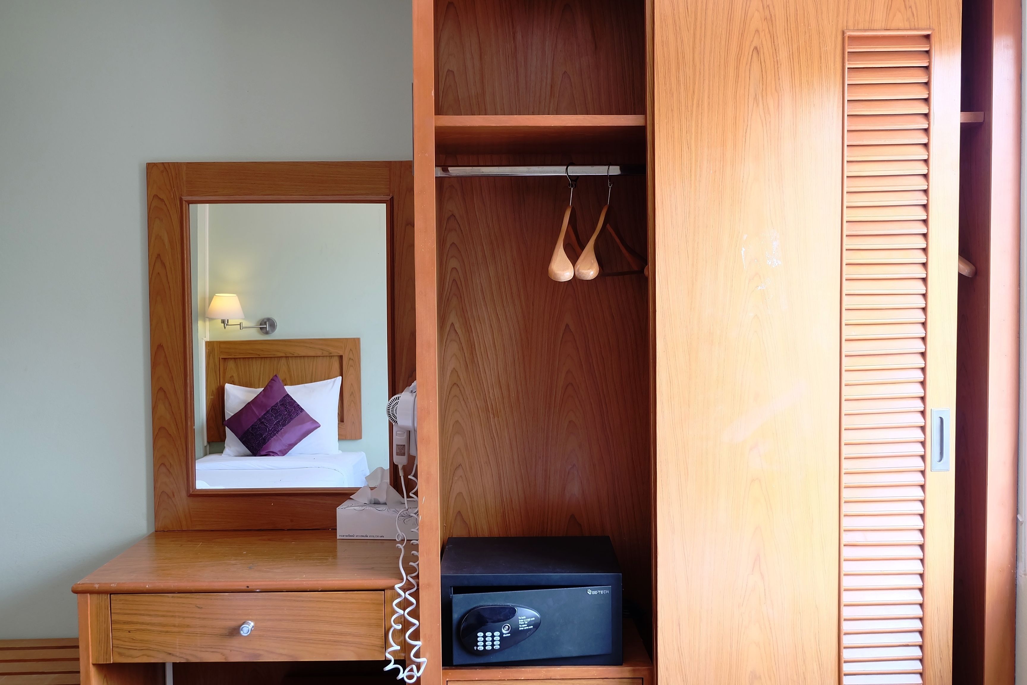 A hotel room with a wooden desk, a bedside table with a lamp, a black safe, and a wardrobe with a mirror.