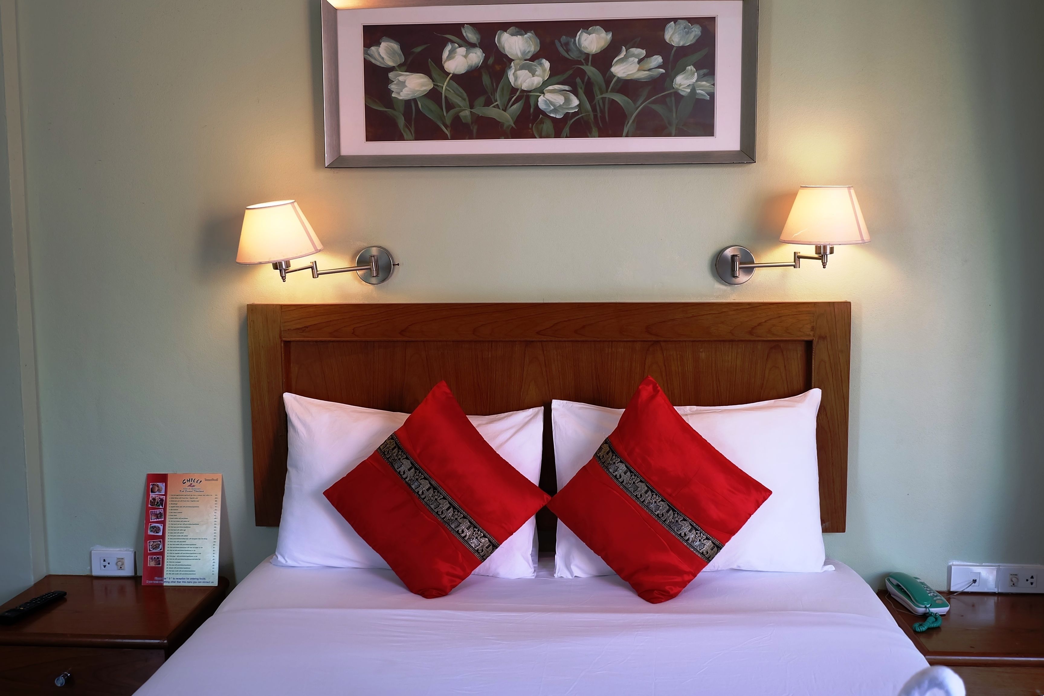 Ein Hotelzimmer mit hölzernem Kopfteil, zwei roten und weißen Kissen, einem gerahmten Blumenbild über dem Bett und zwei Wandlampen.