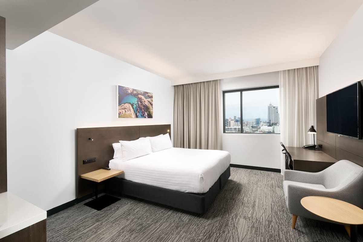 A hotel room with a large bed, a desk, a chair, a TV, and a city view.
