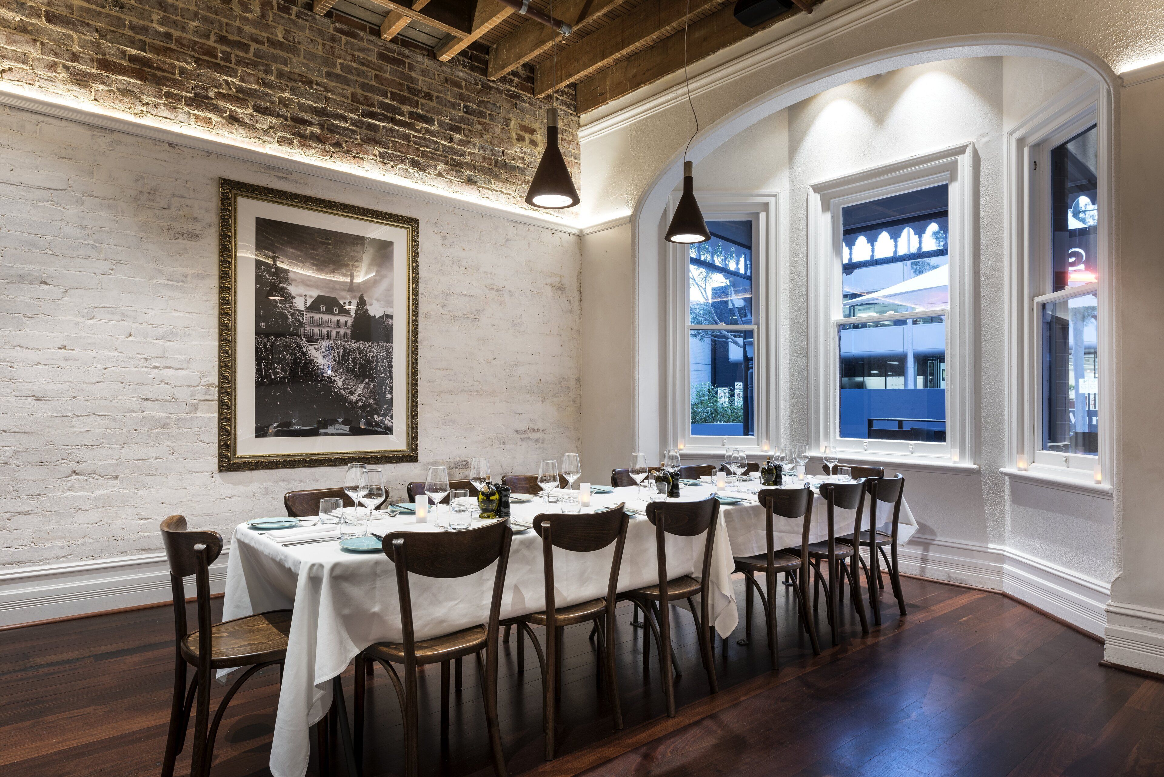 A dining area with a brick wall, a framed picture, and large windows with a view of a blue building.