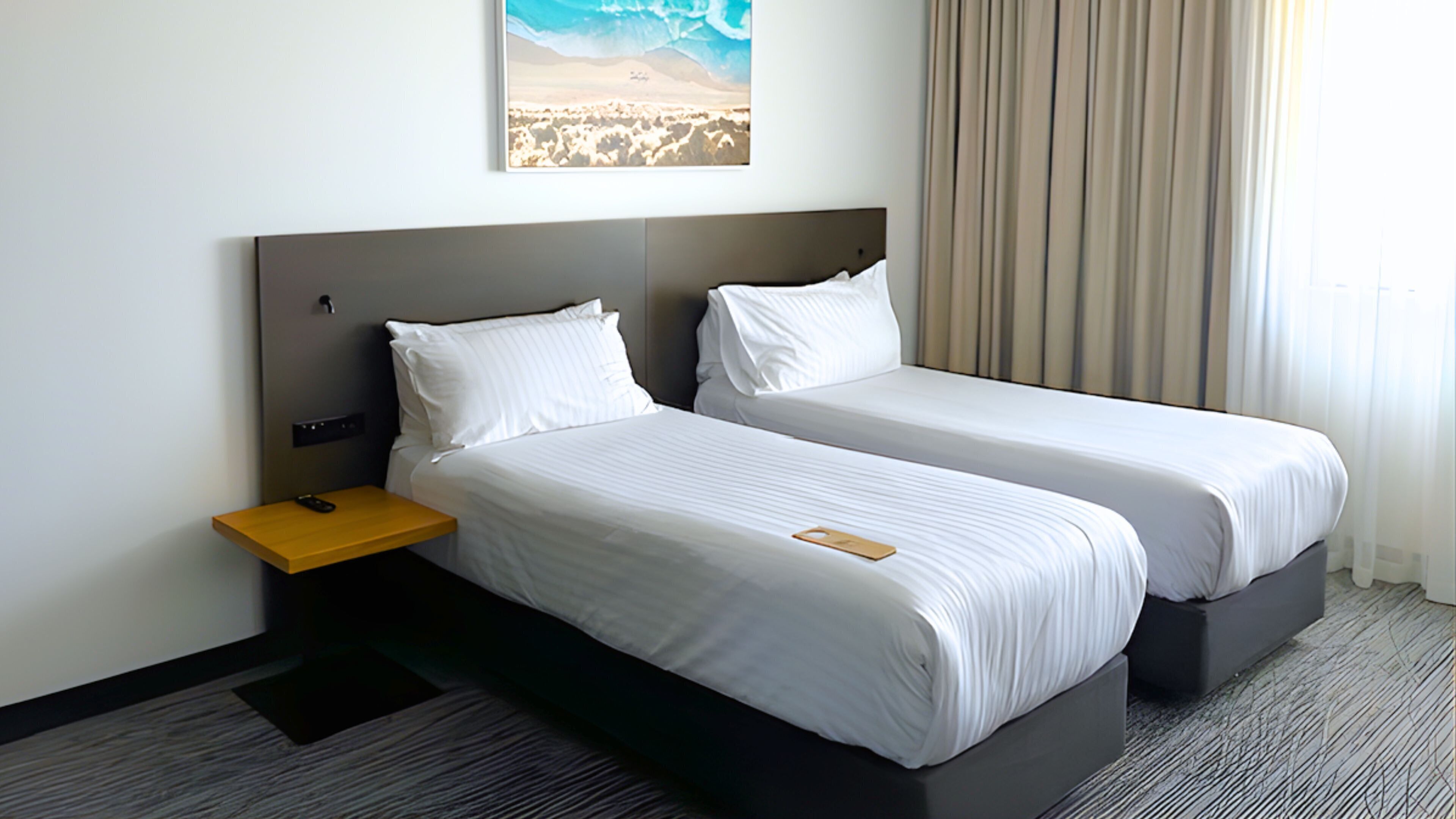 A hotel room with two single beds, a nightstand with a phone, a painting on the wall, and a window with curtains.