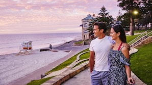 Una pareja parada en una acera con vista a una playa, con una torre de salvavidas y un edificio al fondo.