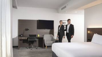 A hotel room with a bed, a desk with a chair, a flat-screen TV, and a small table.
