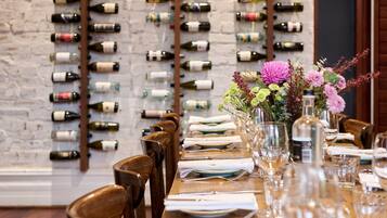 A restaurant with a long wooden table set for dining, a brick wall with wine bottles, and a hanging light fixture.