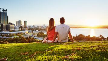A couple sitting on a grassy hill overlooking a cityscape with modern buildings and a body of water during sunset.