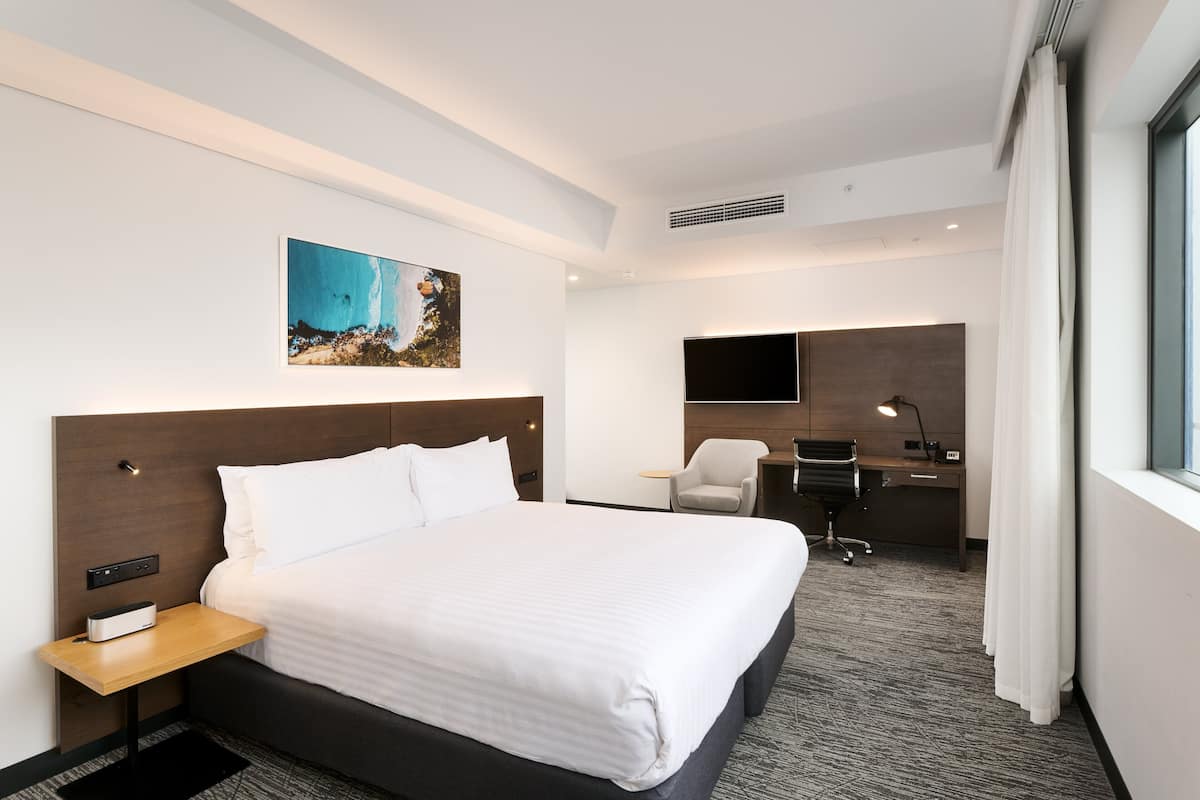 A modern hotel room with a large bed, a desk, a chair, a flat-screen TV, and a painting on the wall.