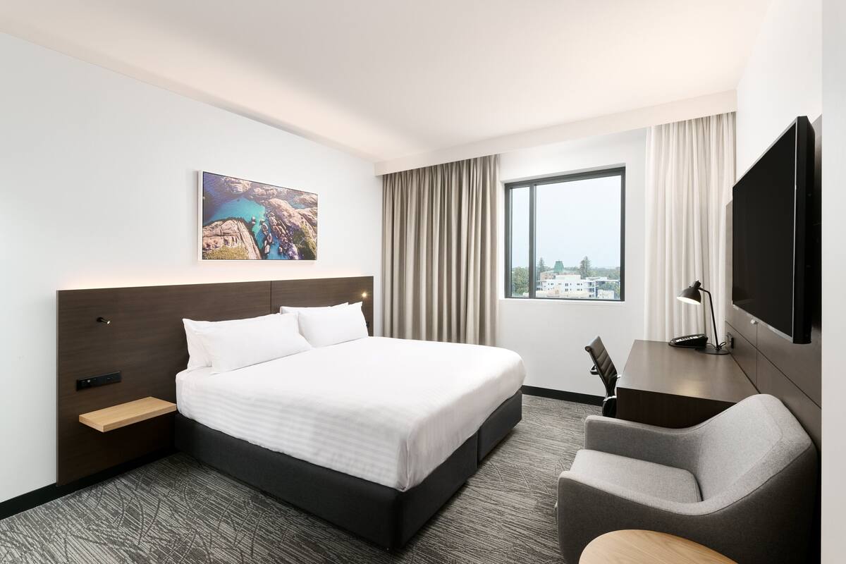 A hotel room with a large bed, a desk, a chair, a TV, a window with curtains, and a painting on the wall.