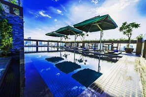 A rooftop infinity pool with lounge chairs and umbrellas, surrounded by stone walls and potted plants.