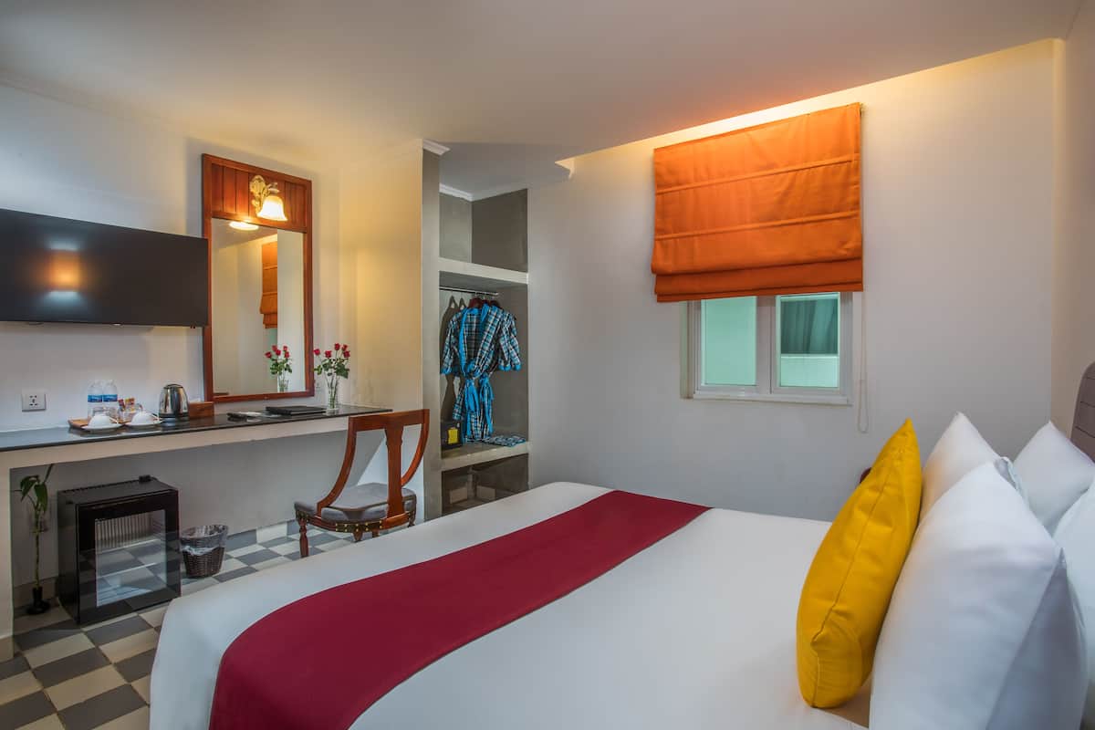 A hotel room with a bed, a desk with a mirror, a chair, a window with a blind, and a small kitchenette area.